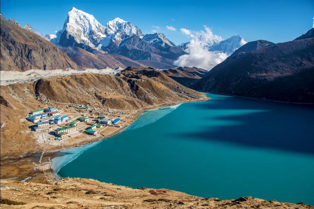 Turquoise alpine lake with mountain lodge settlement surrounded by snow-capped Himalayan peaks and arid valleys