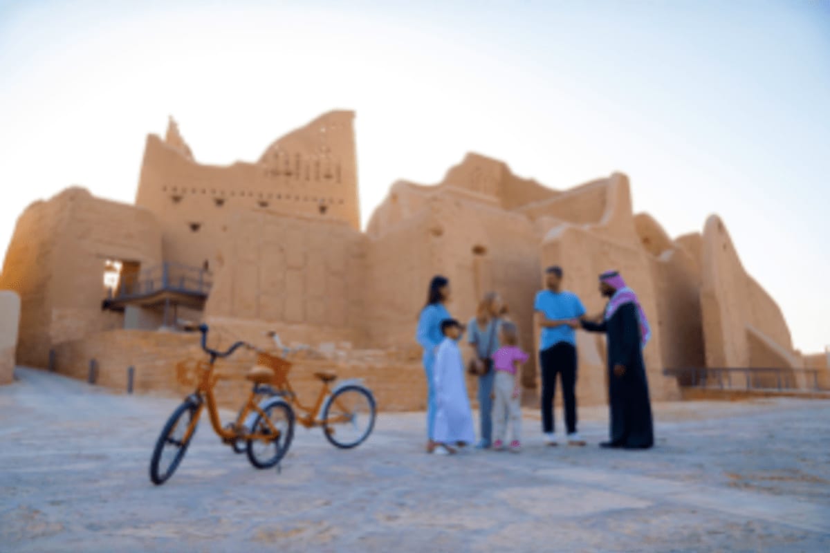Family standing in front of historic mud-brick mosque with yellow bicycle in desert courtyard at sunset
