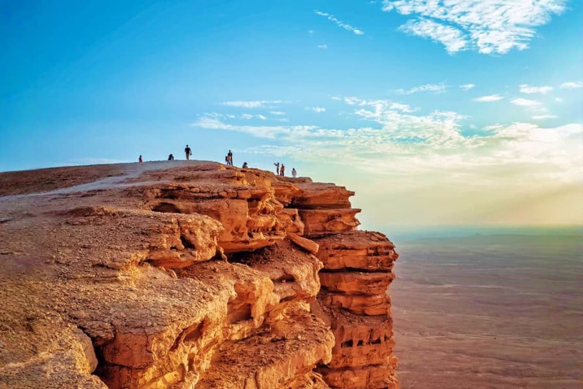 Tourists standing on a rocky cliff at Edge of the World near Riyadh, with vast desert landscape below under a blue sky