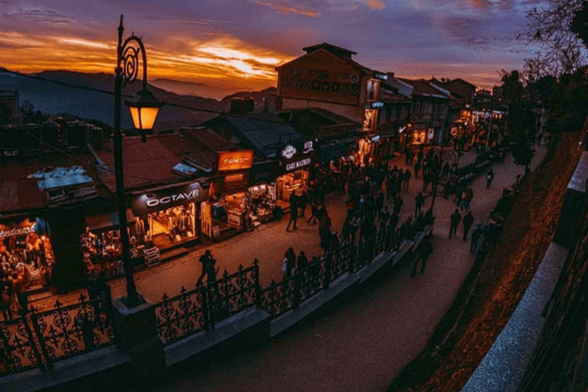 Historic bazaar street at dusk with illuminated shops, ornate railings, and snow-capped mountains at sunset in Himachal Prade