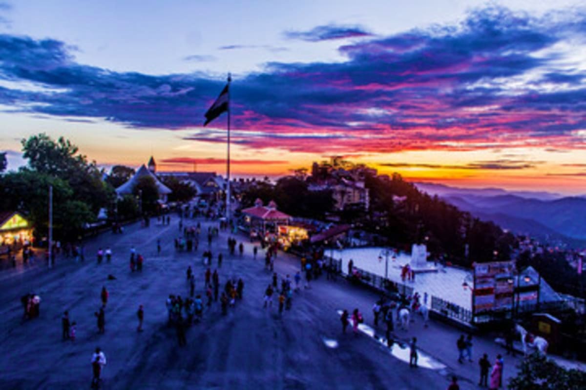 Sunset view of crowded hilltop plaza with flag, buildings, and mountains in Himachal Pradesh hill station