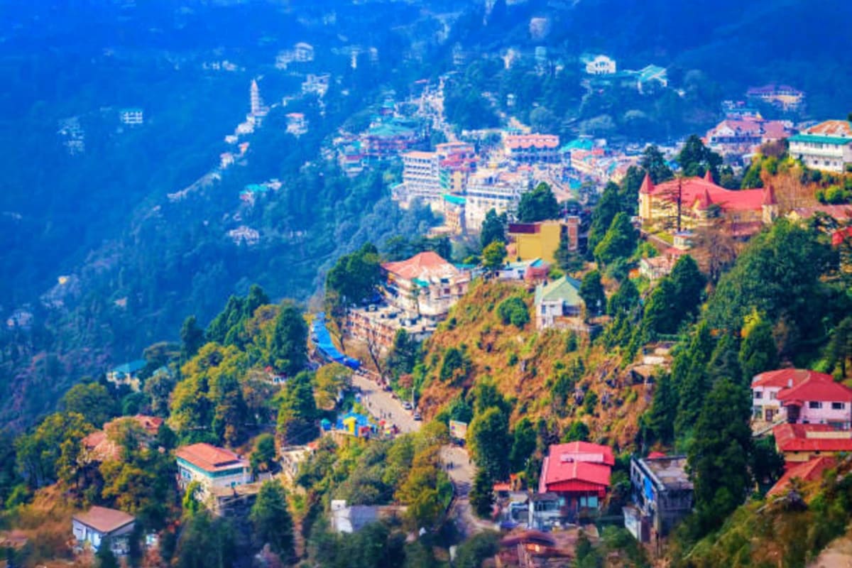Aerial view of Mussoorie hill station with colorful buildings, dense pine forests, and the Doon Valley stretched below under 