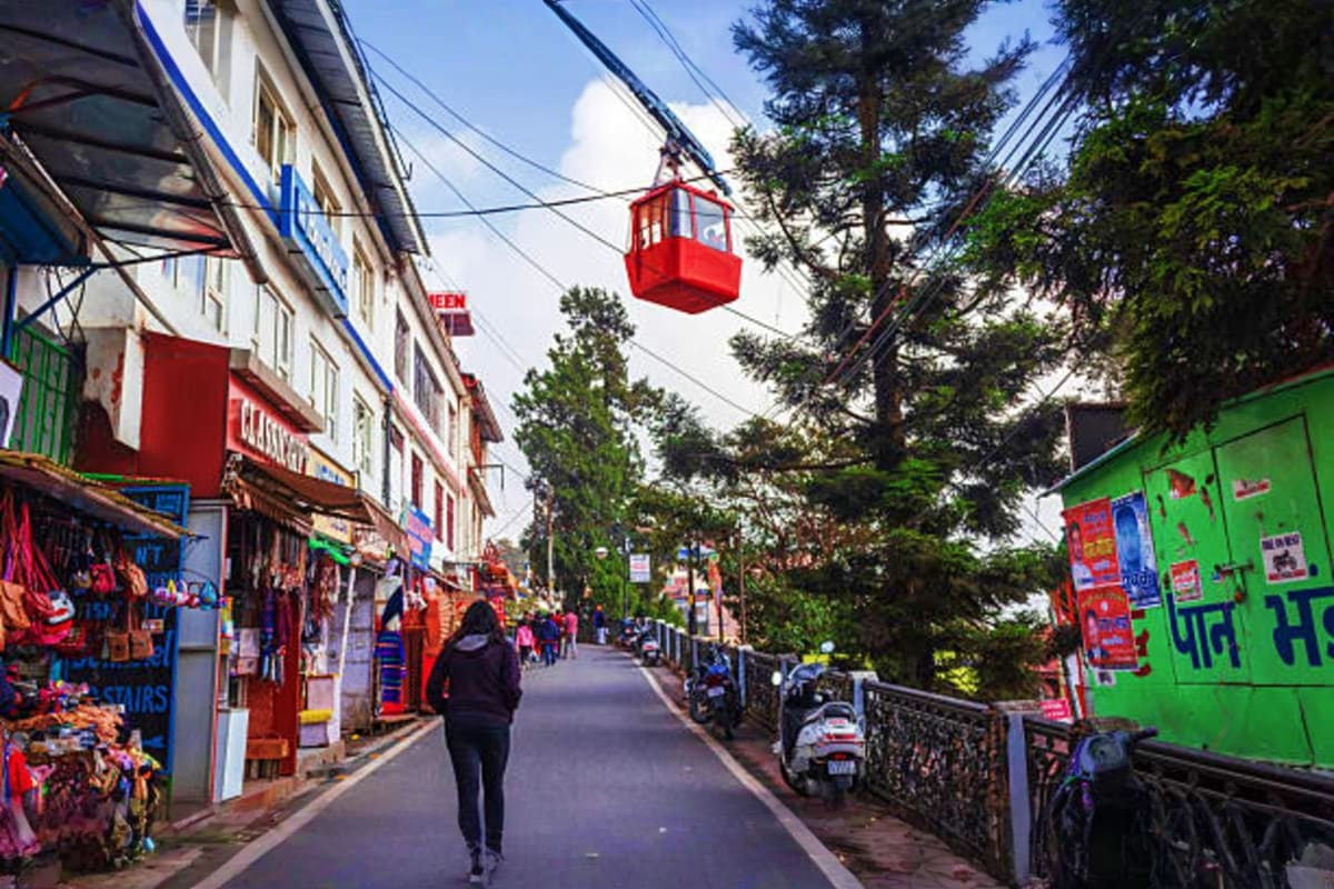 Busy street in Mussoorie with shops, red cable car overhead, tall trees, and pedestrians walking along the commercial area.