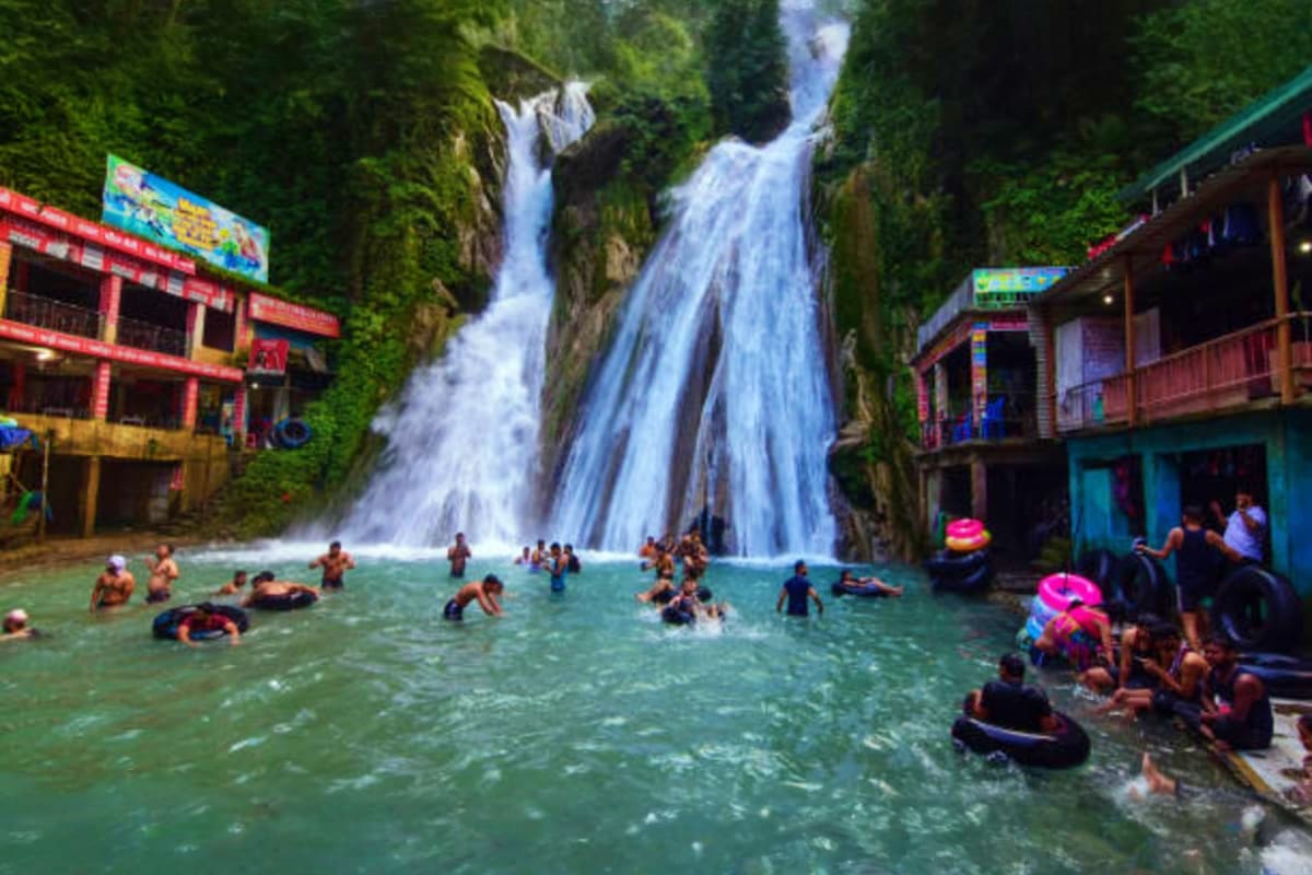 Tourists swimming and enjoying a large cascading waterfall in an emerald pool surrounded by moss-covered cliffs and colorful 