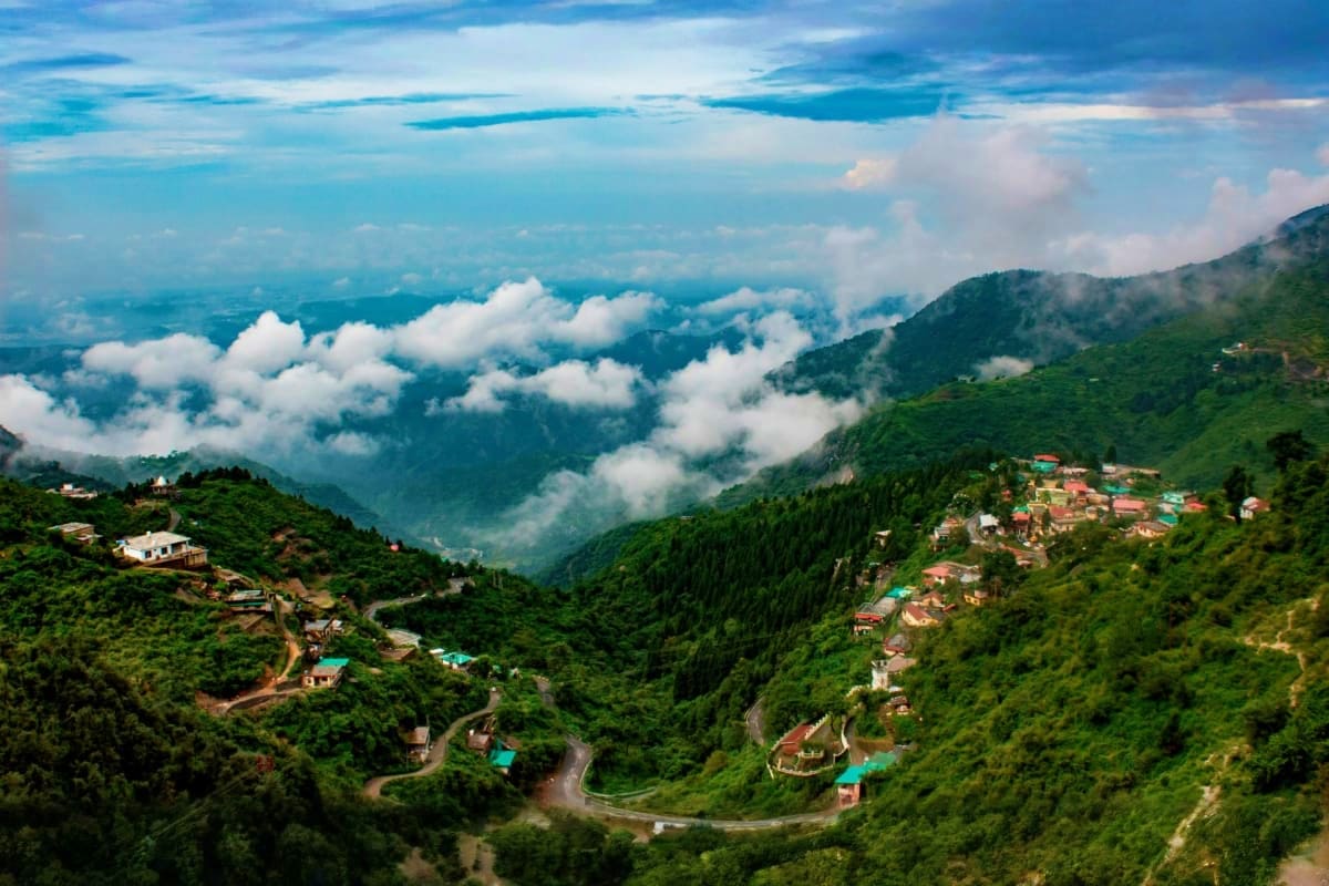 Aerial view of Mussoorie hillside town with winding roads, green forests, scattered houses, and white clouds over distant val