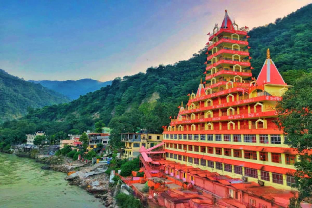 Colorful red and yellow Hindu temple with multiple tiers overlooking the Ganges River in Haridwar, surrounded by forested Him