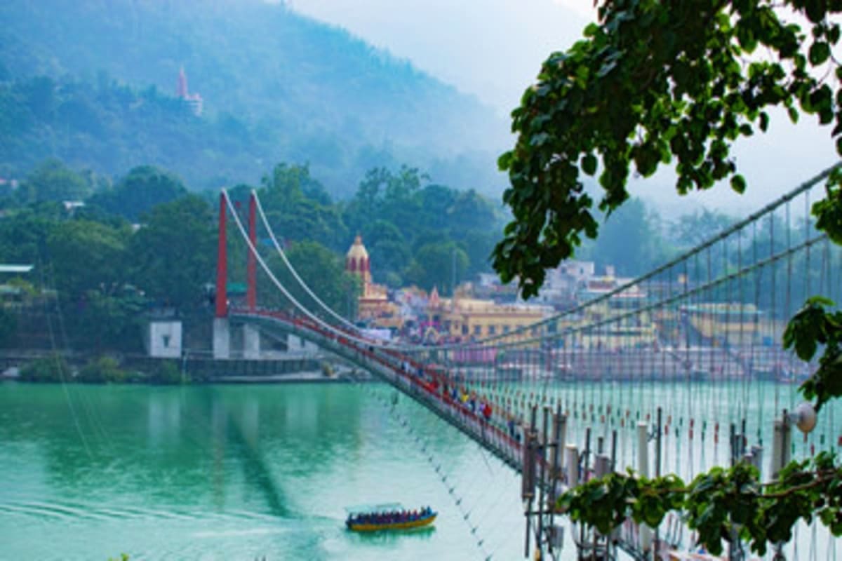 Suspension bridge spanning turquoise river with temple and forested Himalayan mountains in background, boat on water below