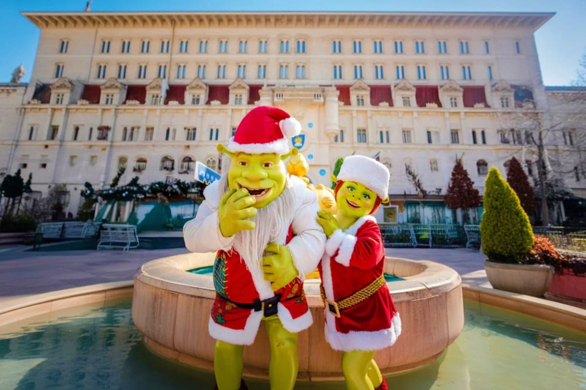Shrek and Fiona characters in Santa outfits posing in front of a fountain at a luxury hotel during Christmas celebrations in