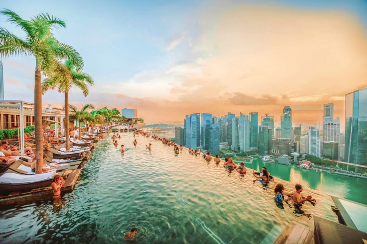 Rooftop infinity pool overlooking Marina Bay with swimmers, palm trees, and Singapore skyline at sunset