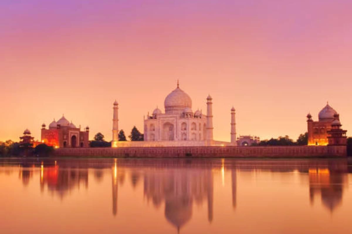 Taj Mahal reflecting in calm water at sunset with pink and orange sky, flanked by smaller domed structures along the riverban
