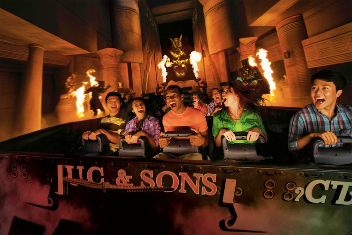 Excited guests on a themed roller coaster ride with fire effects and Egyptian-inspired scenery at an amusement park attractio