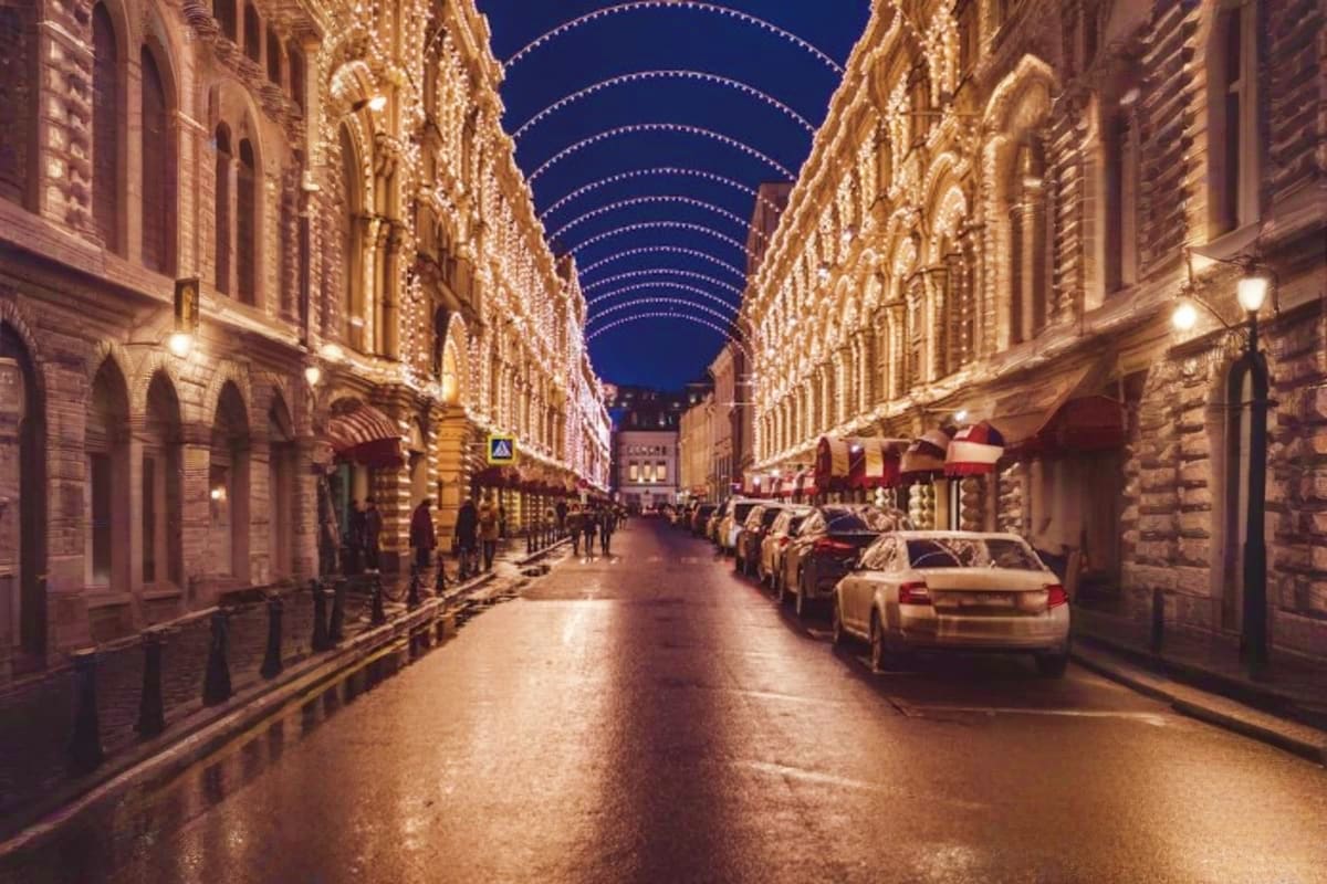 Illuminated Moscow street at night with classical architecture, arched holiday lights overhead, parked cars, and pedestrians 