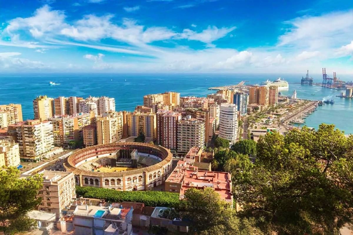 Aerial view of Málaga cityscape with bullring, Mediterranean Sea, port cranes, and high-rise buildings under blue sky