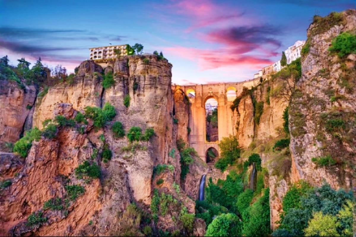 Ronda Spain historic bridge at sunset with dramatic cliffs, illuminated stone arches spanning deep gorge, buildings perched o