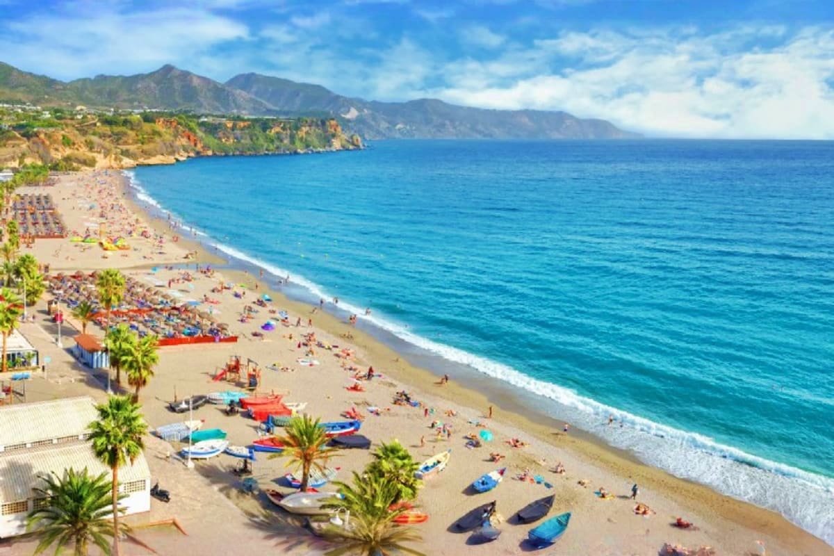 Scenic Mediterranean beach with colorful boats, white umbrellas, palm trees, and dramatic cliffside mountains under blue sky