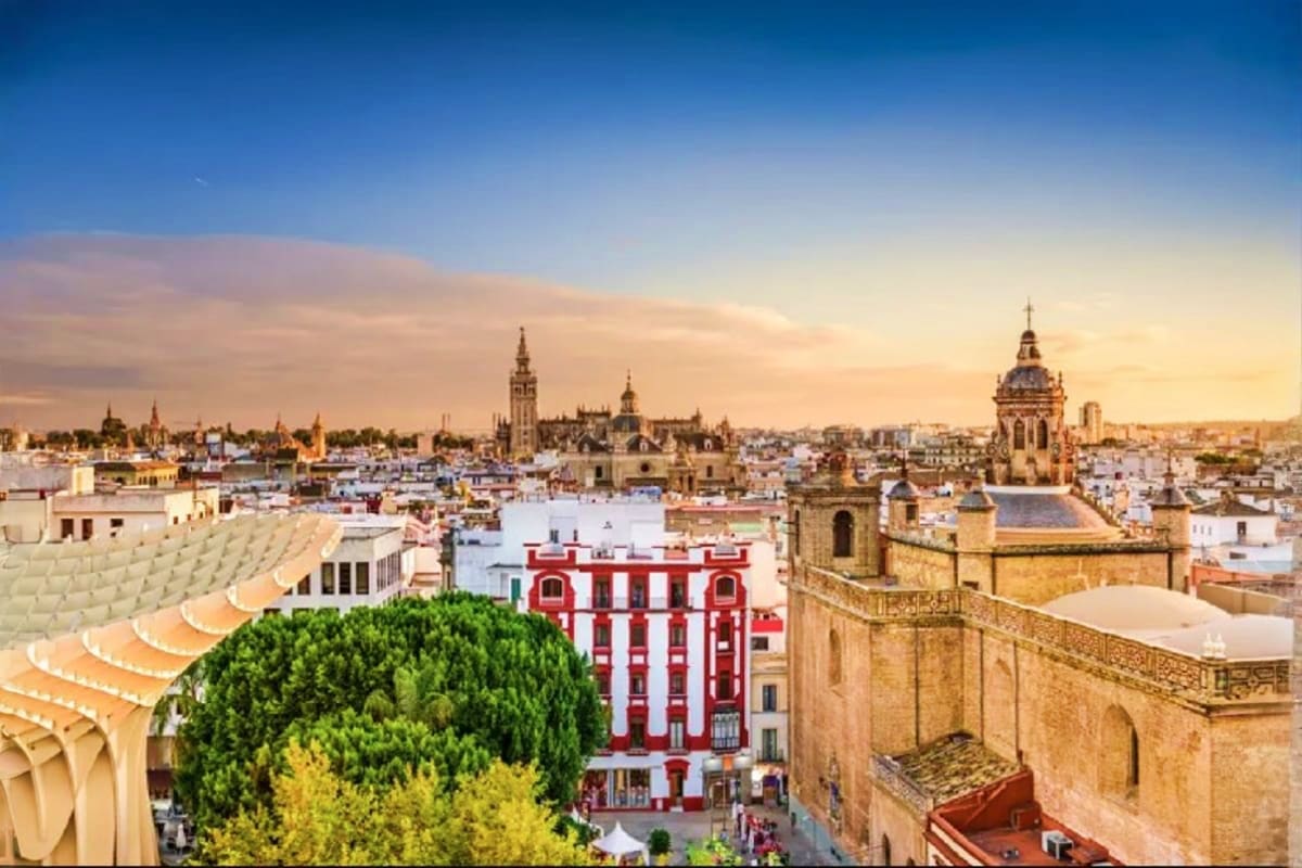 Aerial sunset view of Seville's historic cityscape showing the Cathedral, red colonial buildings, and tree-lined plazas