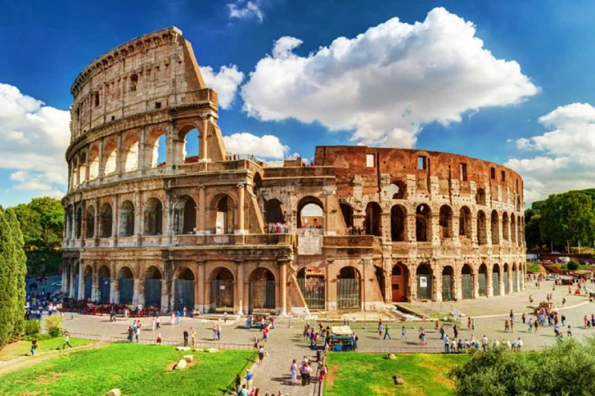 The Essential of Italy The Colosseum in Rome with tourists in the plaza, green lawn, blue sky and white clouds
