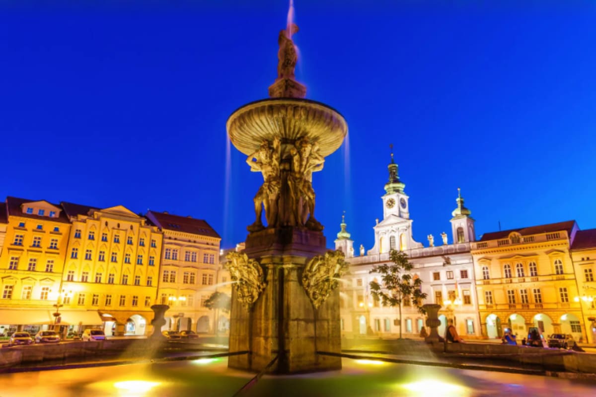Illuminated fountain in historic town square at dusk with yellow baroque buildings and white church with spires in background