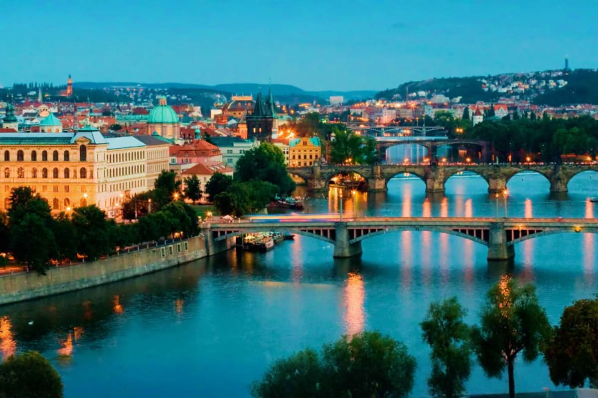 Prague skyline at dusk with Vltava River and illuminated historic bridges, churches, and buildings reflecting in water