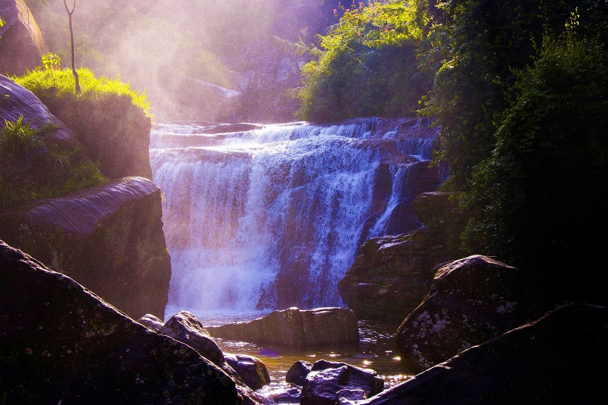 Misty waterfall cascading through lush green forest canyon with sunlit mist and rocky boulders, typical of Sri Lanka's natura