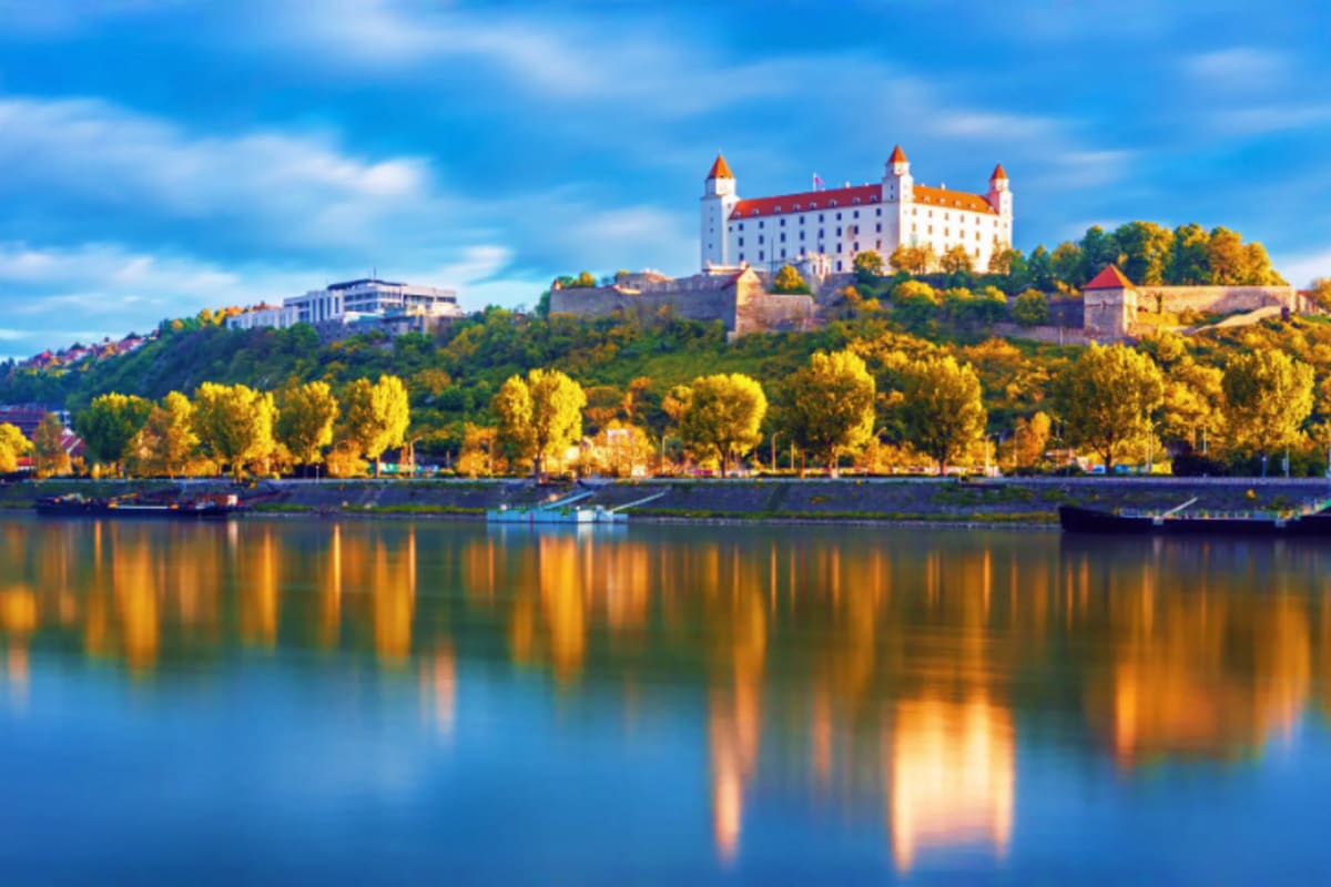 Bratislava Castle with red-roofed towers reflected in the Danube River during autumn with golden foliage