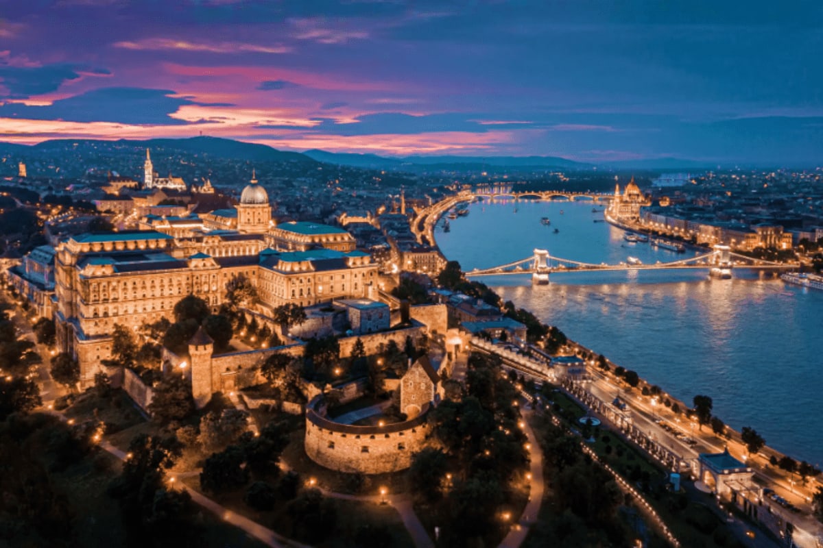 Aerial view of Budapest at dusk showing Buda Castle, Danube River, Chain Bridge, and illuminated cityscape with hills in back