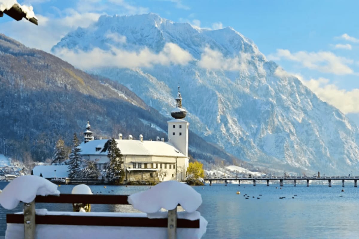 Snow-covered chapel with white tower on lake island, snow-capped Alpine mountains and forested slopes in background, winter l