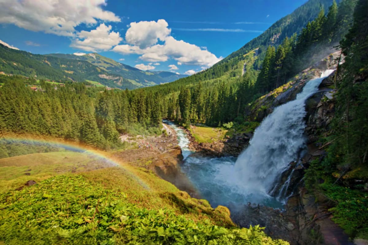 Waterfall cascading down rocky cliff in Alpine valley with coniferous forest, river, and mountain peaks under blue sky with w