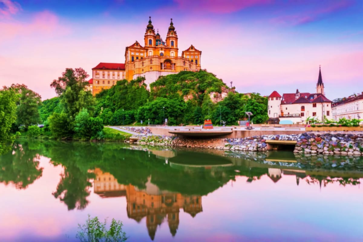Orange baroque monastery church perched on hilltop overlooking Danube River with reflection in calm water at sunset, Austria