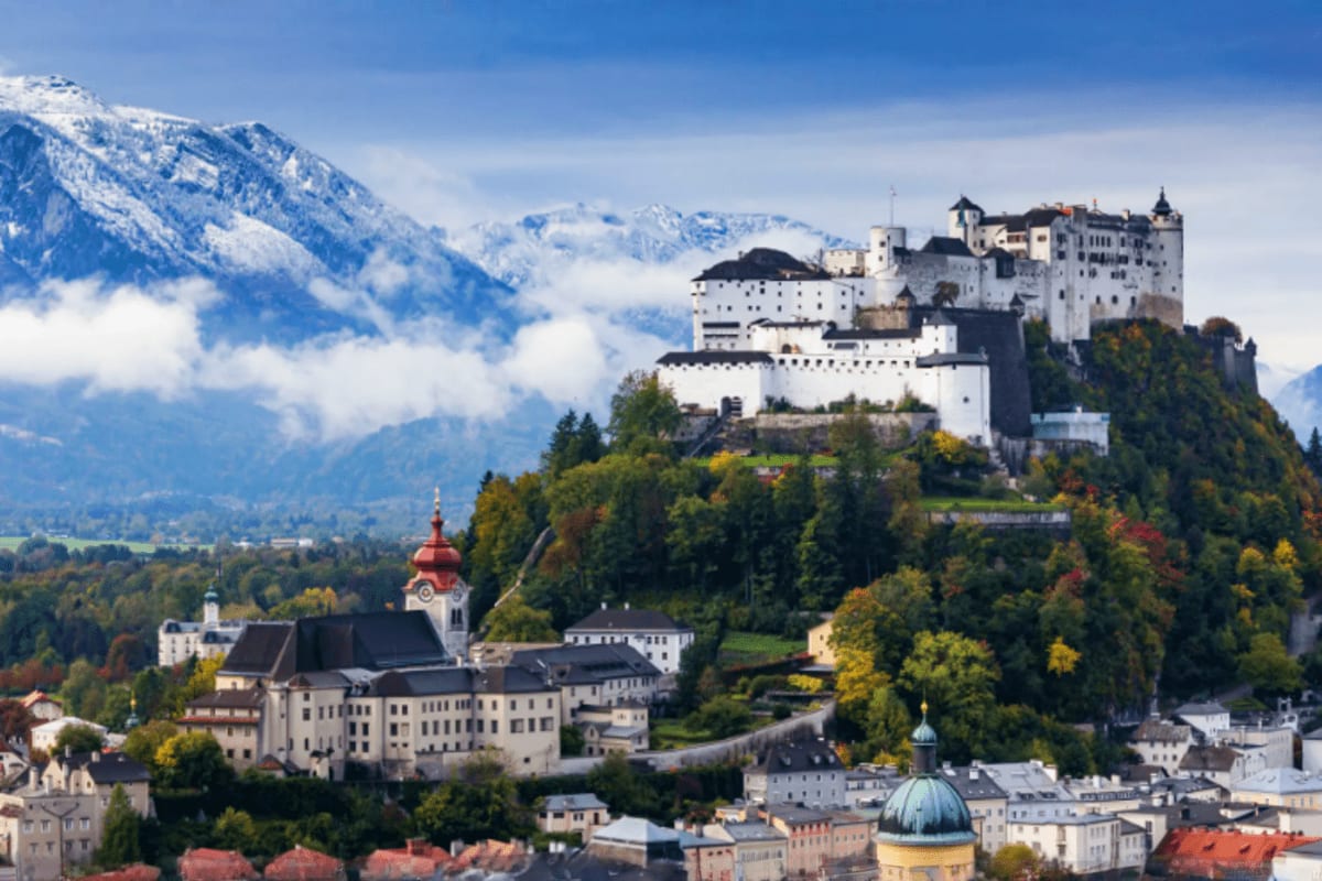 Hohensalzburg Castle perched on a hilltop overlooking Salzburg with snow-capped Alps in the background and a church with red 