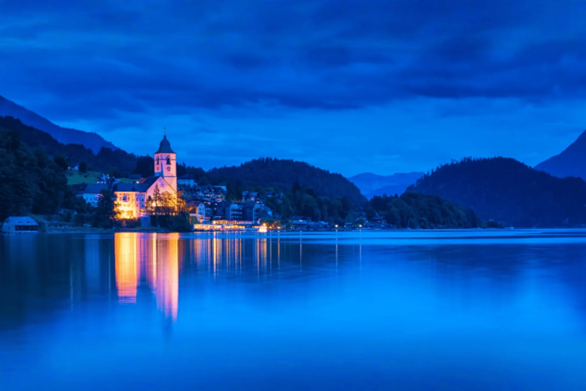 Illuminated church with golden spire reflecting in calm blue lake at twilight, surrounded by forested mountains in Austrian A