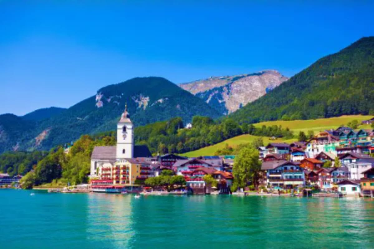 Alpine lakeside village with white church steeple, traditional chalets, turquoise water, and forested mountains under blue sk