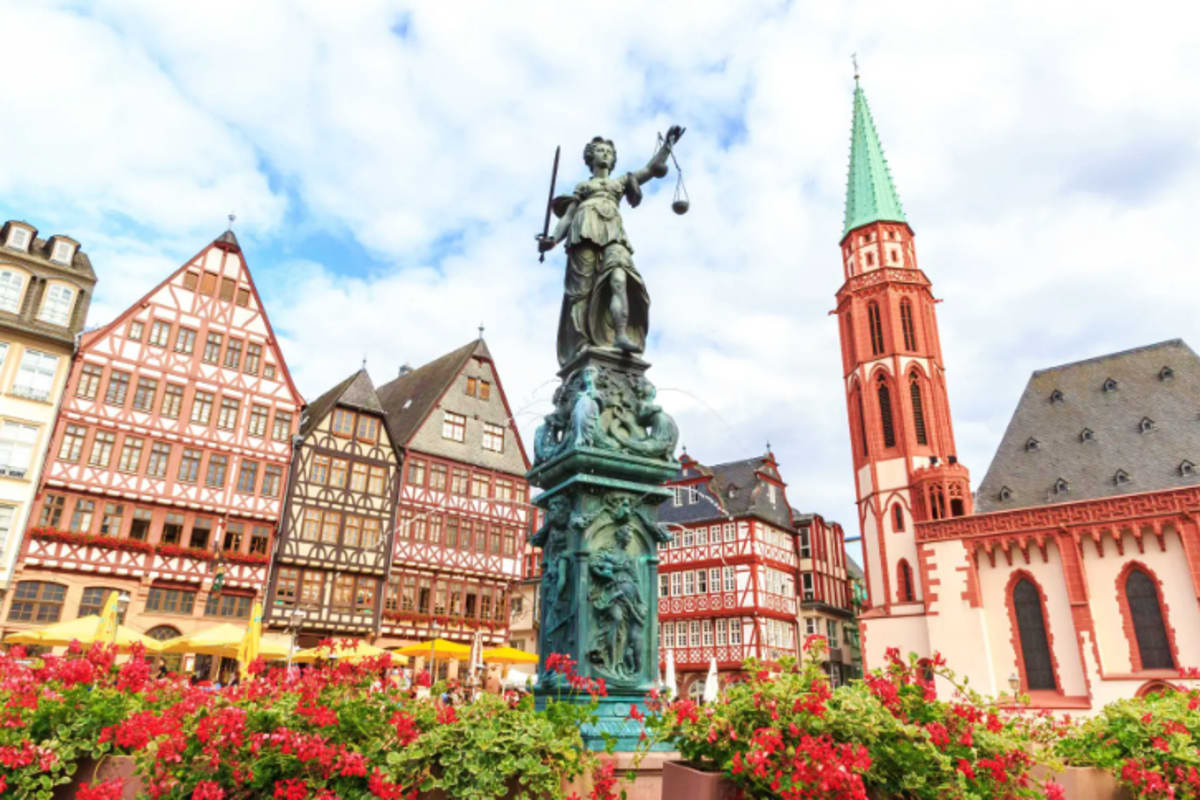 Statue of Lady Justice in Frankfurt's Romerberg square surrounded by red flowers and traditional half-timbered buildings with