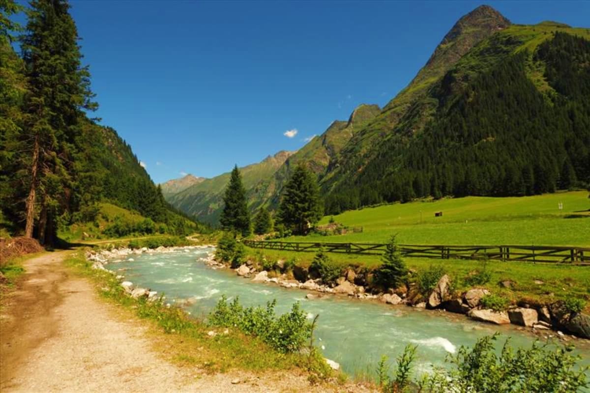 Alpine valley with turquoise river, coniferous forests, meadow, wooden fence, and snow-capped mountains under clear blue sky
