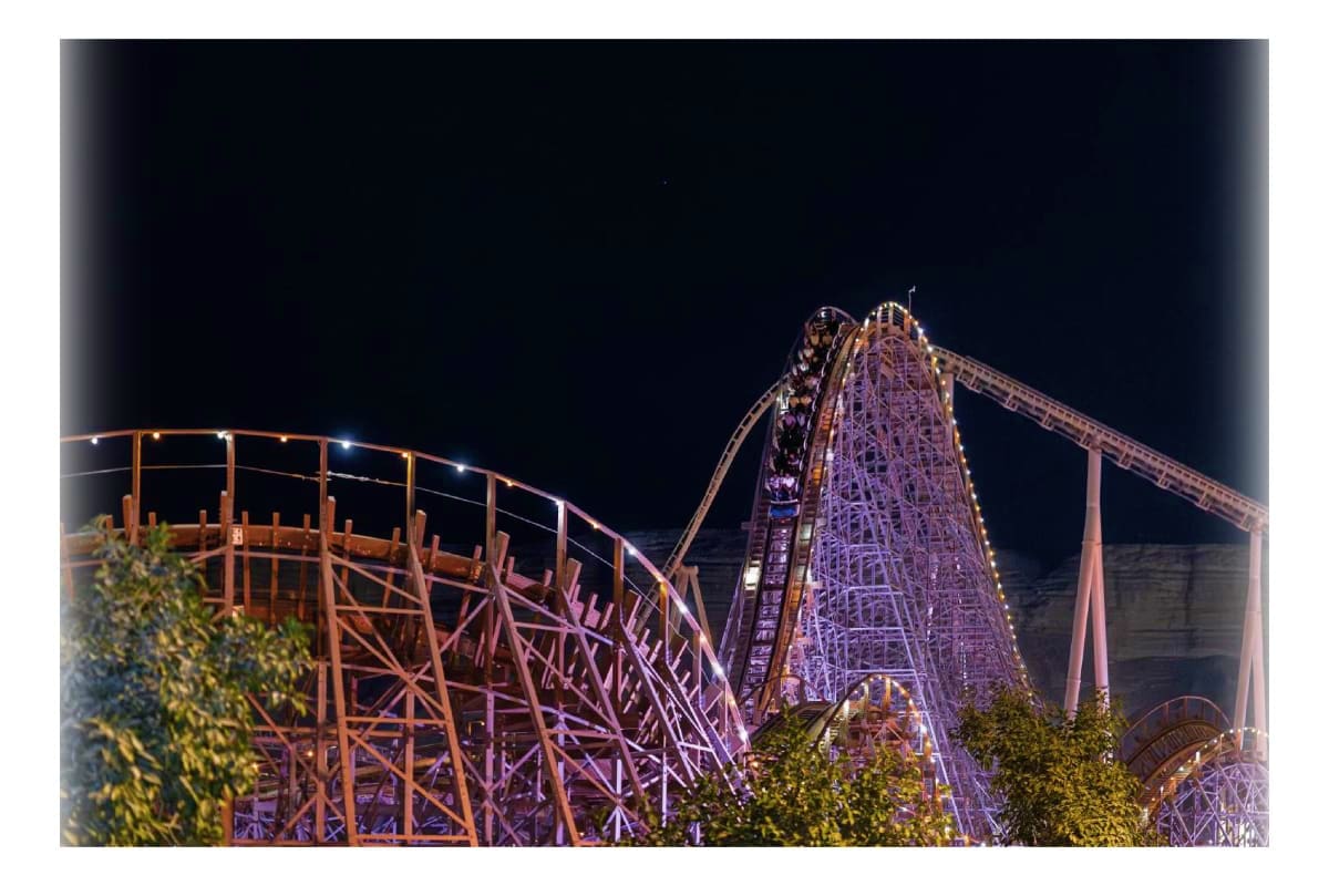 Illuminated roller coaster at night with riders climbing steep wooden and metal track structure against dark sky