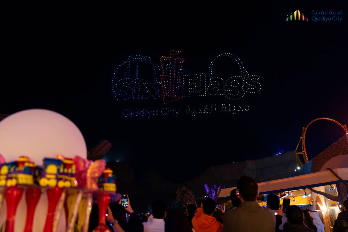 Nighttime drone light show spelling "SIX FLAGS" over Qiddiya City theme park with crowds watching and amusement rides visible