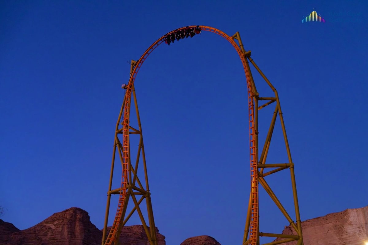 Red and yellow roller coaster loop against clear blue sky at dusk with desert rock formations below