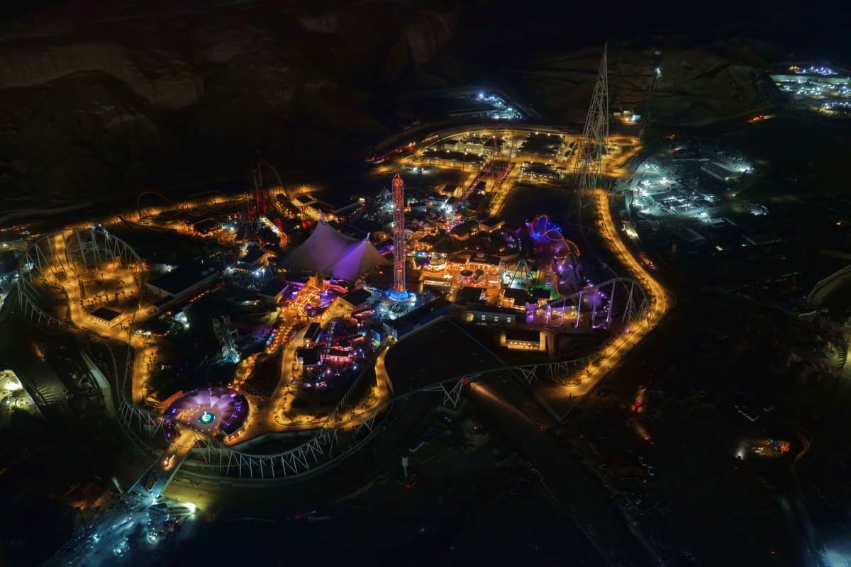 Aerial night view of illuminated amusement park with roller coasters, purple-lit attractions, and tall tower structure during