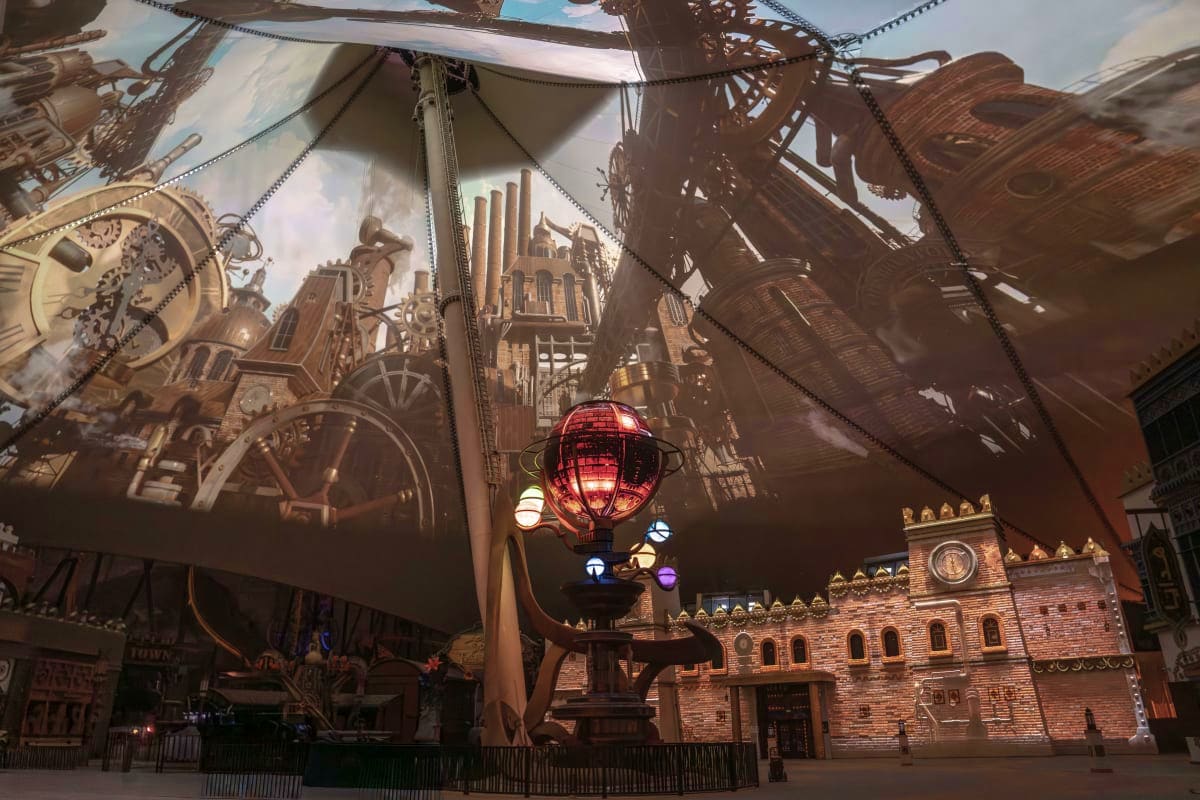 Steampunk-themed indoor theme park attraction with illuminated red airship and ornate brass machinery and buildings displayed