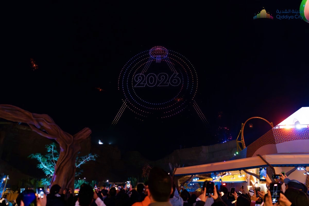 Drone light show displaying "2028" in the night sky above a crowded outdoor event with illuminated structures and carousel vi