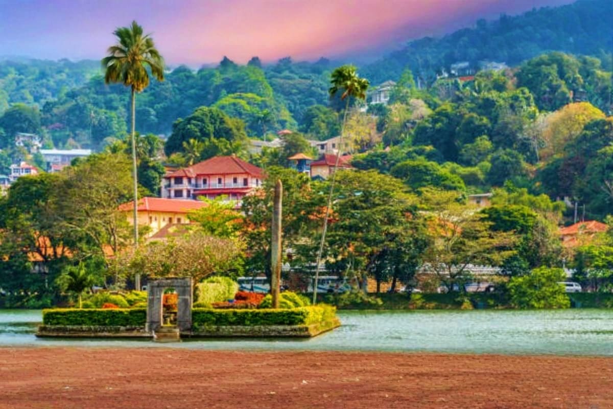 Scenic view of Kandy Lake with colonial architecture, palm trees, and lush green mountains at sunset in Sri Lanka