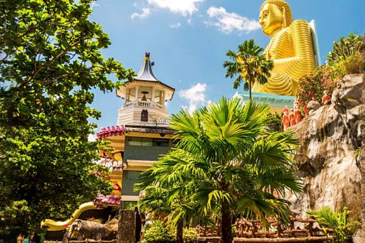 Large golden Buddha statue at a Sri Lankan temple with white bell tower, palm trees, and stone monastery buildings under blue