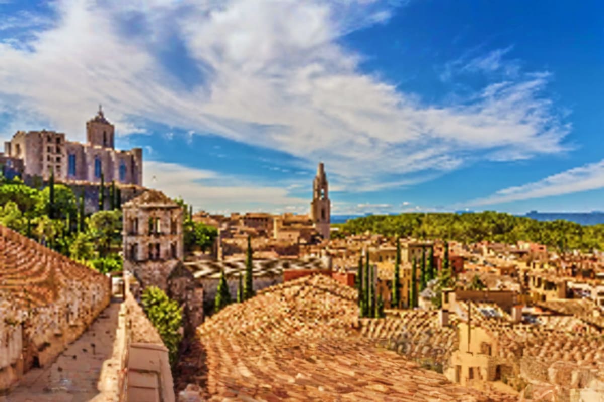 Medieval hilltop town with stone buildings, church tower, and cypress trees overlooking Mediterranean landscape under blue sk
