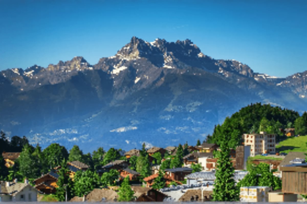 Snow-capped Alpine mountain peak with traditional chalets and green trees in Swiss valley landscape under clear blue sky