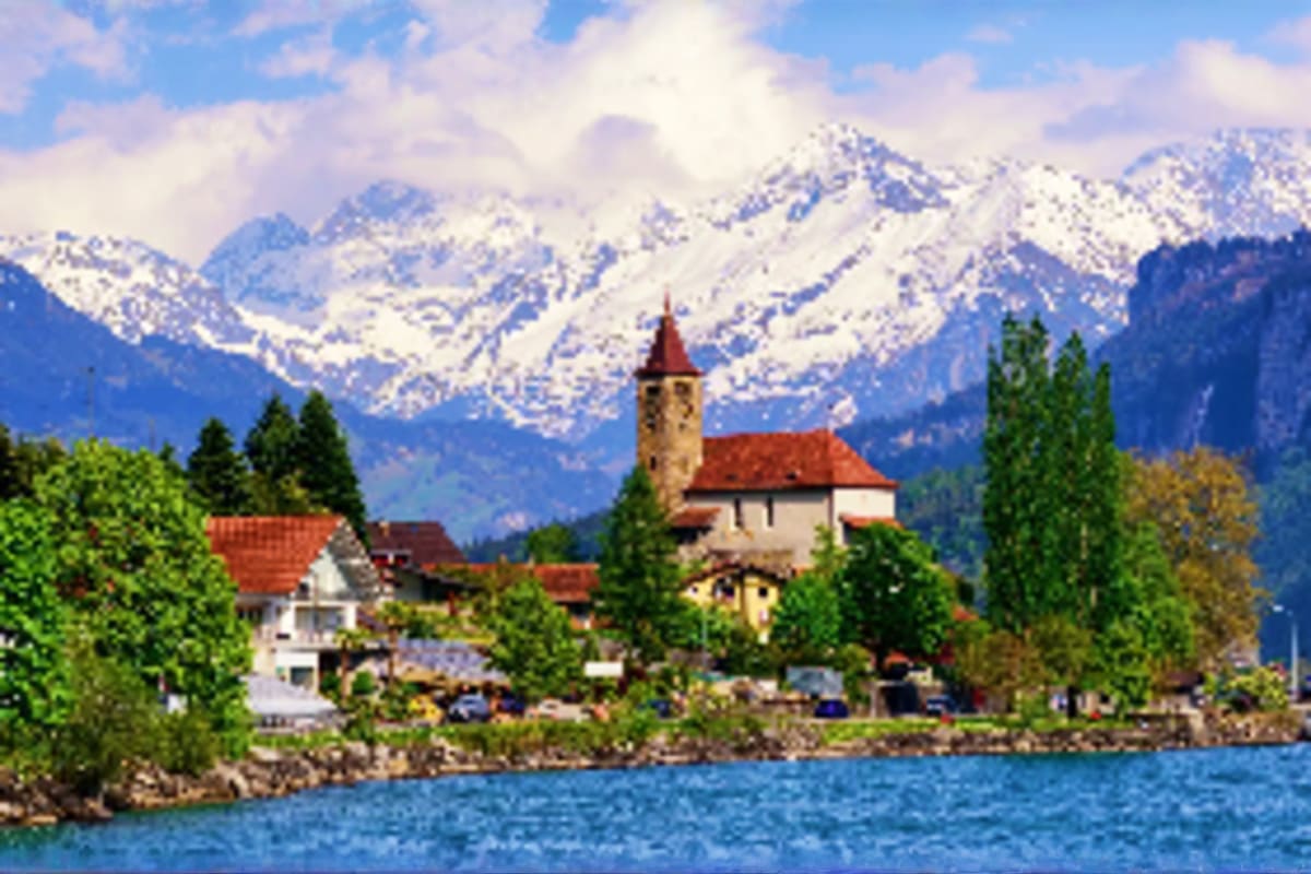 Alpine village with church and bell tower on lakeside, snow-capped mountains in background, evergreen trees and traditional E