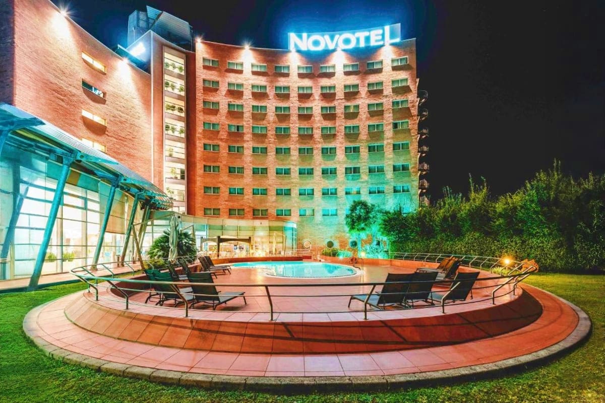 Novotel hotel at night with illuminated brick facade, outdoor pool surrounded by lounge chairs, and manicured grounds