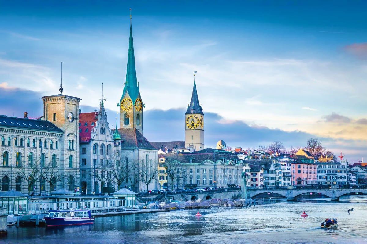 Zurich waterfront with historic churches, green spires, and colorful buildings reflected in the Limmat River under blue sky