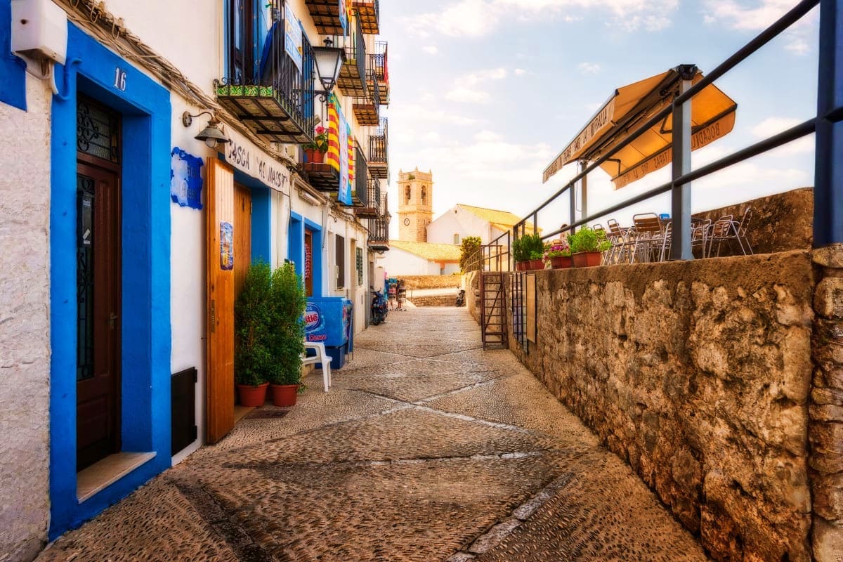 Narrow cobblestone street in historic Spanish town with blue and white buildings, stone walls, and church tower in background