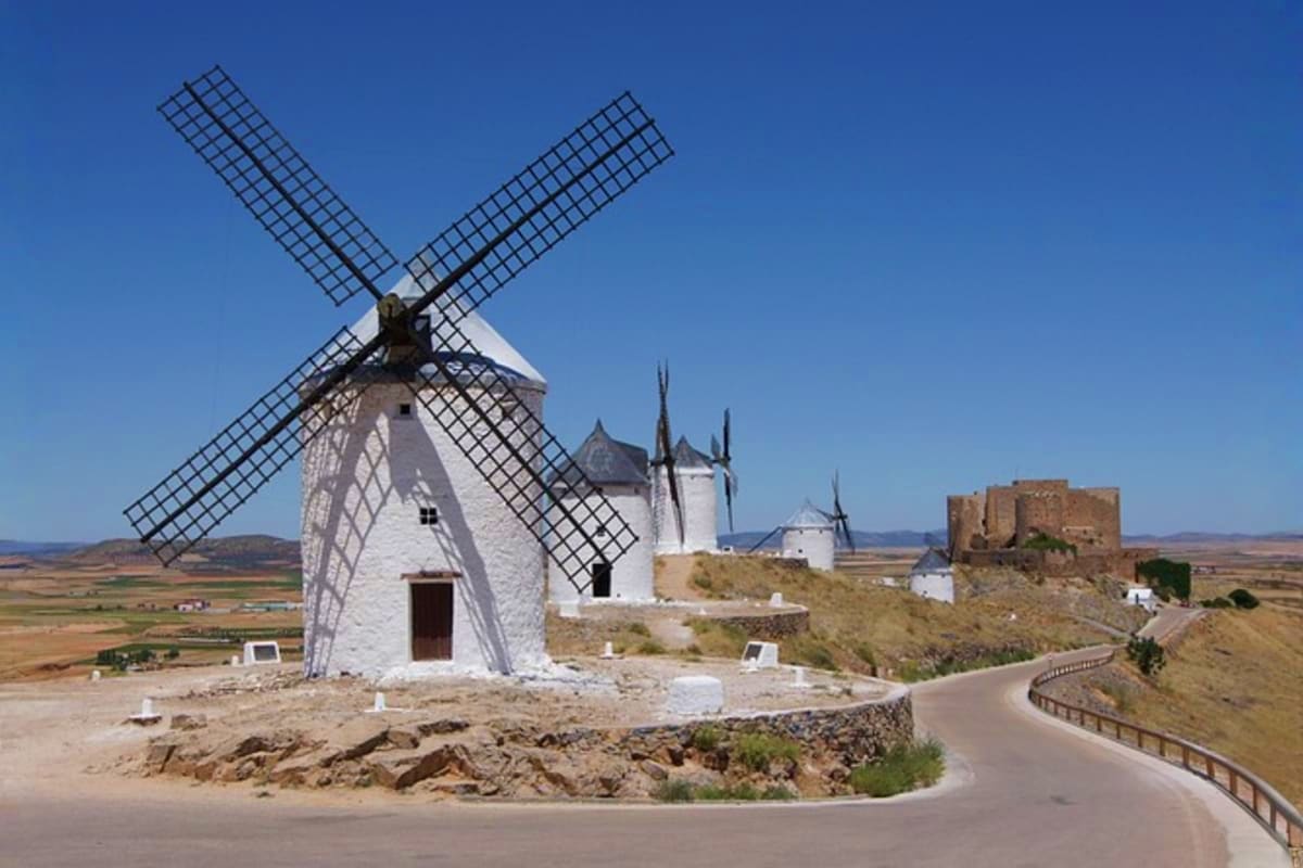 White windmills with latticed sails on hilltop in La Mancha, Spain, with castle ruins visible in distance under clear blue sk