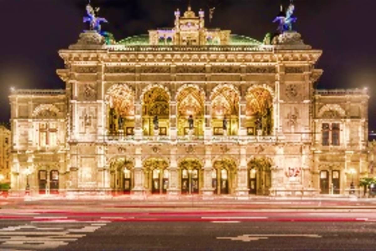 Ornate beige stone opera house facade with golden arched windows lit up at night, with decorative sculptures on roof.